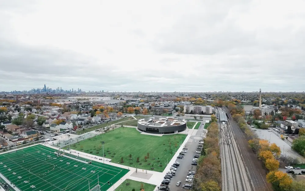 High aerial over sports fields, rail lines and autumn trees, with a distant city skyline on the horizon.
