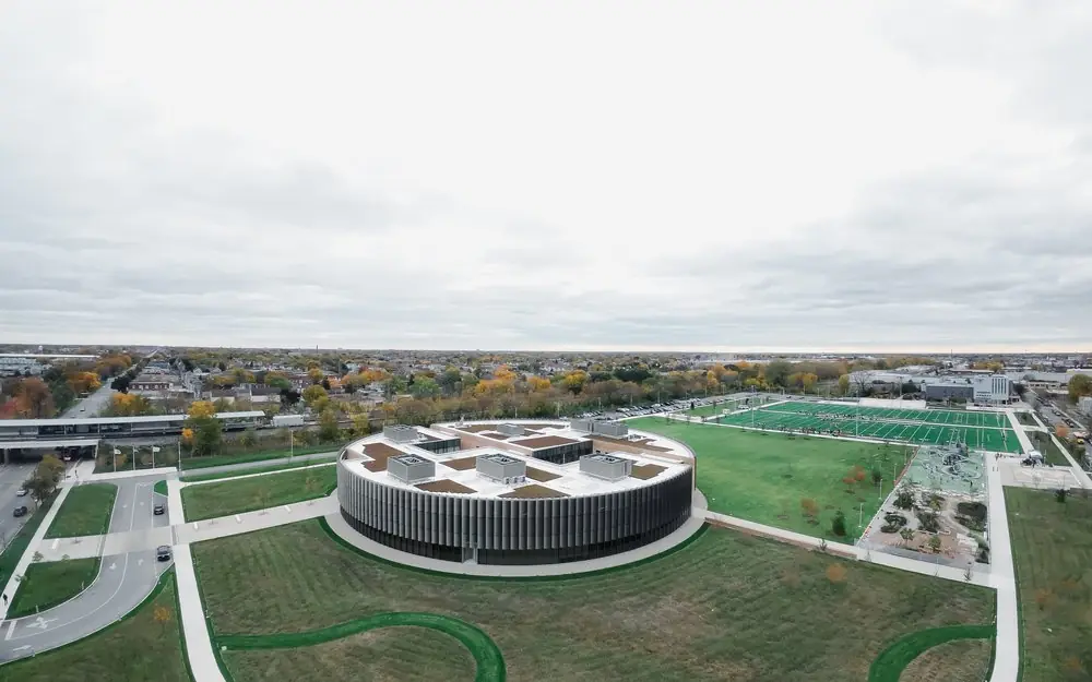 Wide aerial of a circular building, green fields and a running track on an autumn afternoon.
