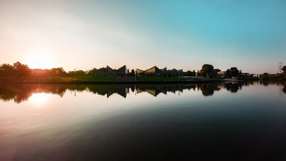Low sun over still water at golden hour, a row of peaked rooftops and trees mirrored in the surface.