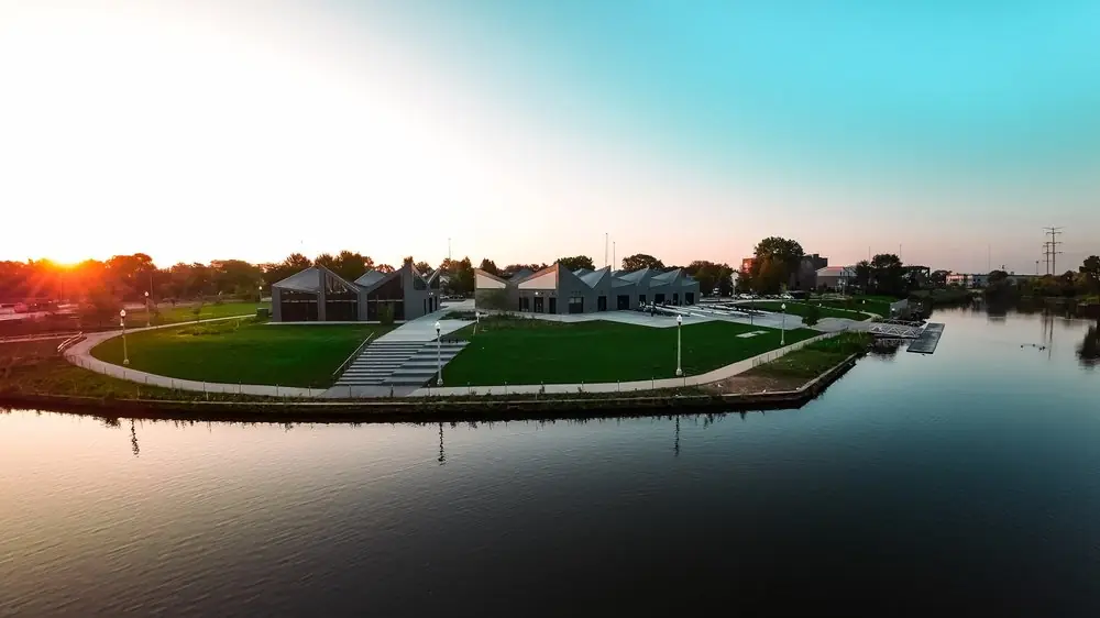 Drone shot at sunset of a waterfront park with angular rooftops, a curved path and calm water.