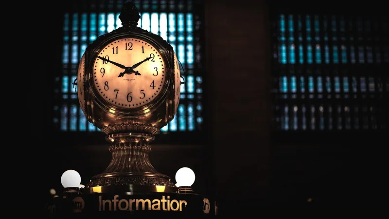 A glowing brass four-faced clock above an information booth, set against tall dark windows at night.