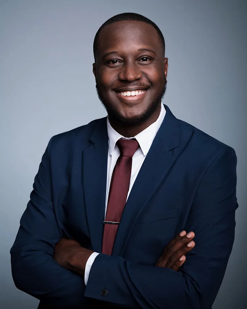 Portrait of Ruona Ebiai, smiling with arms folded, in a navy suit and burgundy tie.